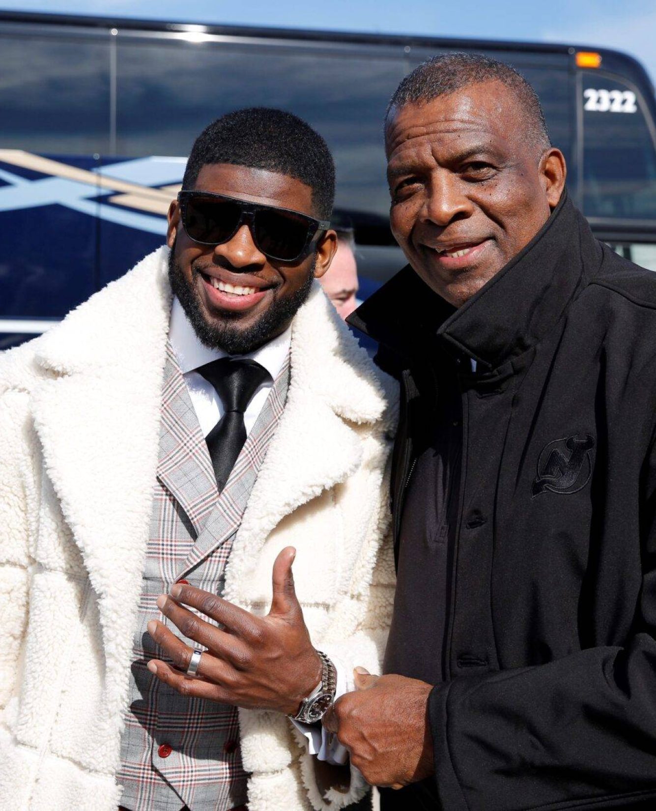 Two Men, One In A White Fur Coat And Sunglasses, And Another In A Black Jacket, Stand Smiling In Front Of A Bus.